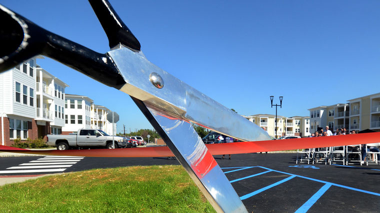 Scissors are cutting a ribbon with people and building with parking on the background.